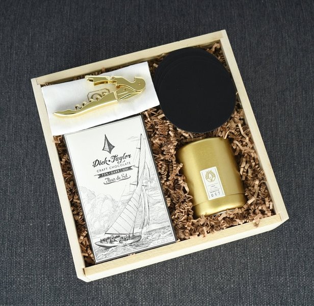 A wooden Occasions Box crate packed with a gold candle, a gold corkscrew, craft chocolate and slate coasters, on charcoal linen