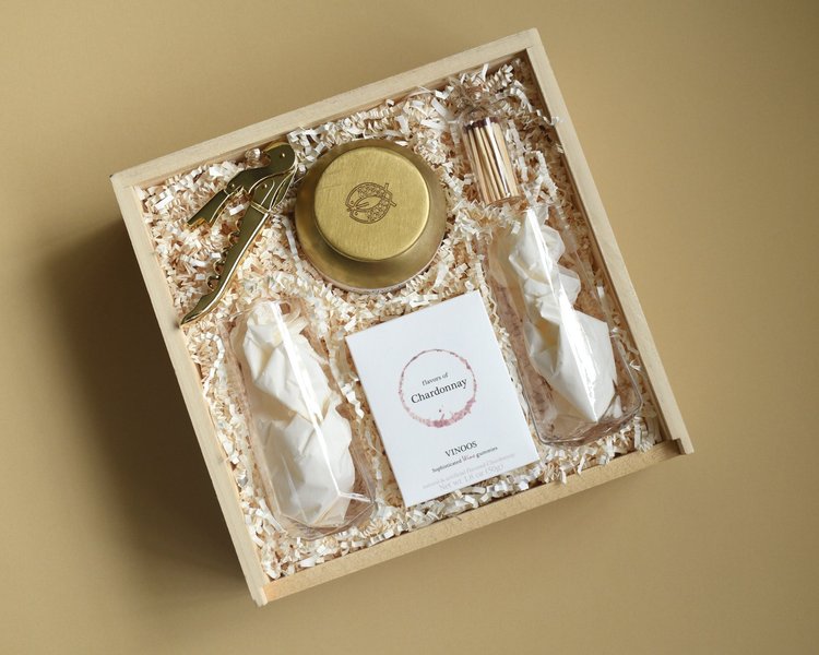 A wooden crate holding two crystal wine glasses, a gold candle, a gold corkscrew and a box of chardonnay gummies
