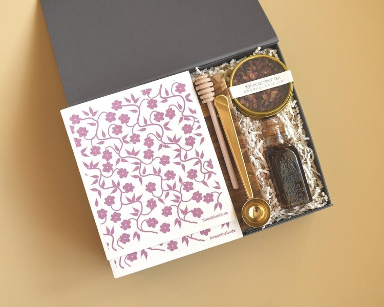 A charcoal box holding rose mint loose-leaf tea, a honey jar and dipper, a gold spoon and floral-print dishcloths