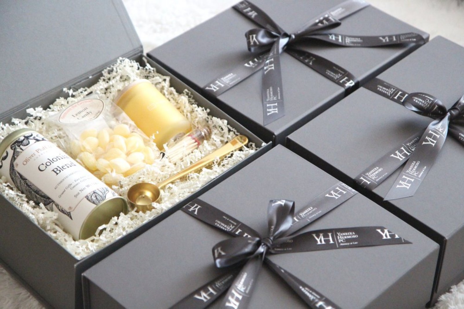 Four signature charcoal Occasions Box gift boxes tied with ribbon printed with a client's monogram, one open to show coffee, lemon candy, a candle and a gold scoop