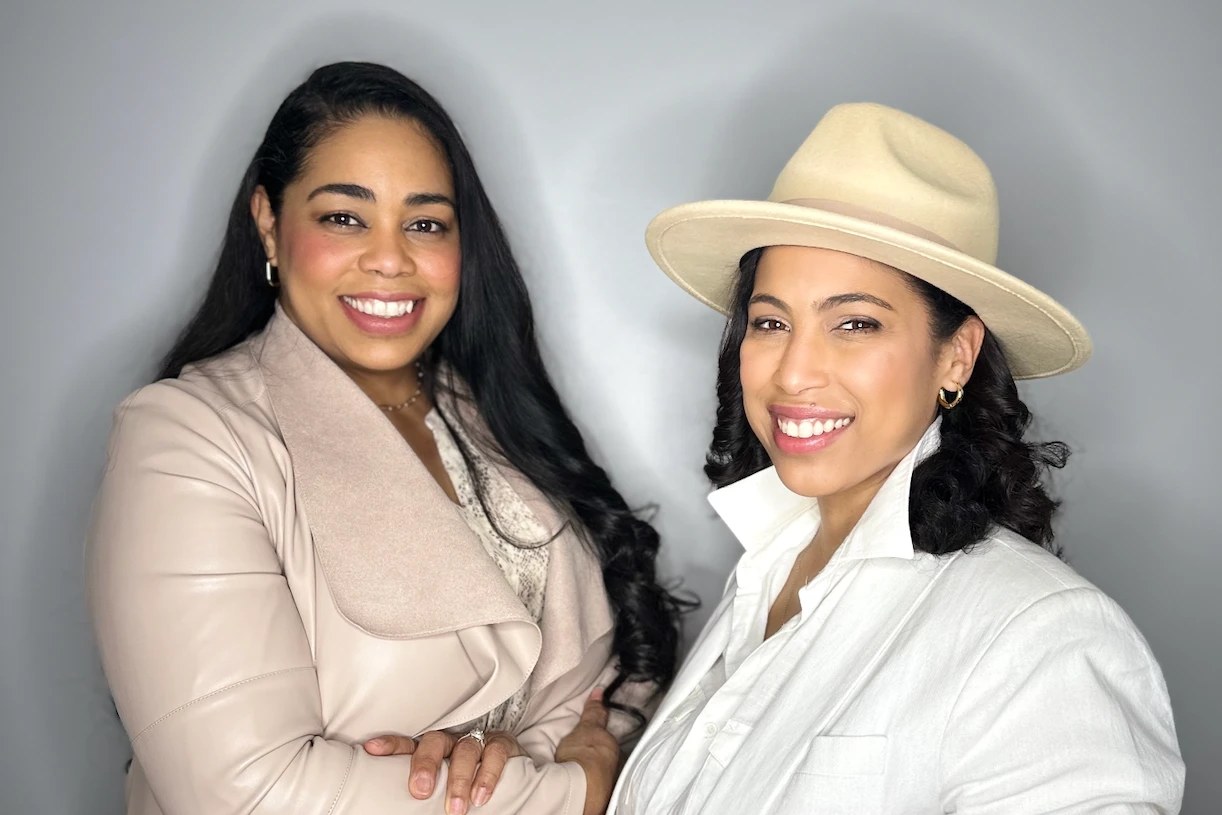 The two sisters who founded Occasions Box, standing side by side against a pale grey wall, one in a blush leather jacket and the other in a white linen shirt and a cream wide-brim hat.