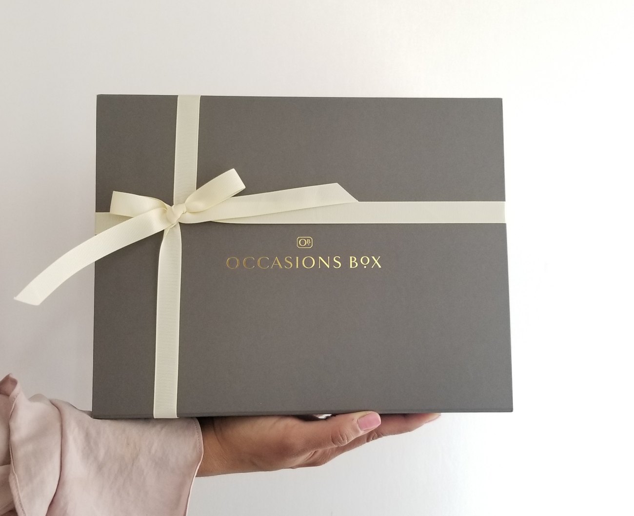 A signature charcoal Occasions Box with a gold embossed lid and ivory ribbon, held in one hand